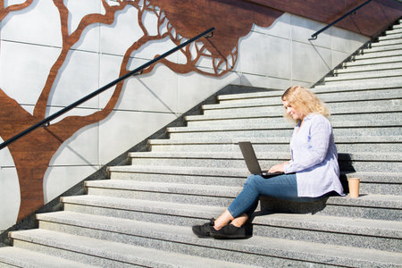 Young blonge woman using her laptop outside. Sitting on stairs, urban styleの写真素材