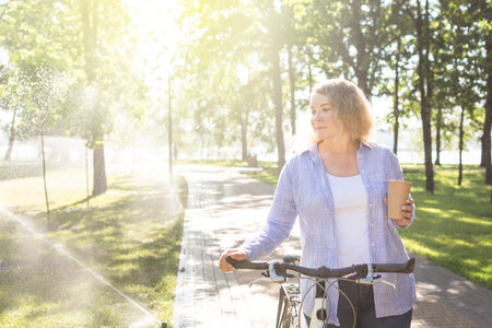 Young blonge woman walking her bicycle in the park and holding a cup of coffeeの写真素材