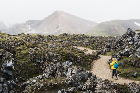 Woman in yellow raincoat walking through lava field towards foggy mountains in Landmannalaugar National Park, Icelandの写真素材