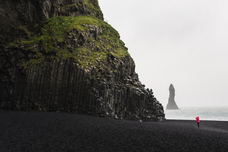 Woman in red raincoat taking pictures of the black basalt columns at the black sand beach in Vik, Iceland.の写真素材