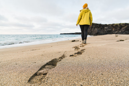 Girl in yellow raincoat walking on the sand of Skardsvik beach at sunset. Focus on footprints, wide angle lens shot, Snaefellsnes peninsula, Iceland.の写真素材