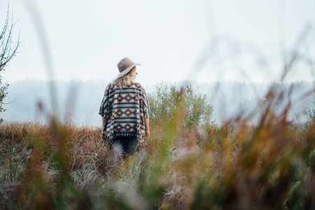 Woman in wide-brimmed felt hat and authentic poncho standing in high brown grass at foggy morning.の写真素材