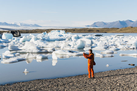 FJALLSARLON, ICELAND - AUGUST 2018: Man in orange jacket taking pictures of floating ice in Vatnajokull glacier lagoon.のeditorial素材