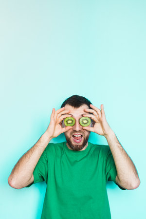 Young bearded holding slices of green kiwi fruit in front of his eyes, mouth wide open. Light green background, vertical orientation, copy space.の写真素材
