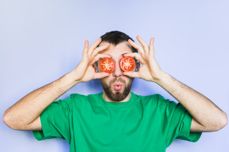 Young bearded man holding slices of red tomatoes in front of his eyes, surprised face expression. Light purple background, copy space.の写真素材