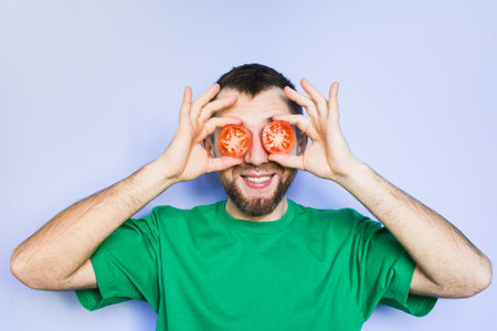 Young bearded and smiling man holding slices of red tomatoes in front of his eyes. Light purple background.の写真素材