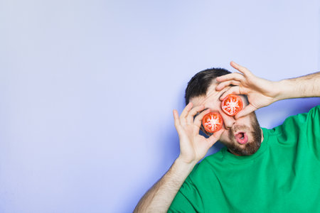 Young bearded and showing surprise man holding slices of red tomatoes in front of his eyes. Light purple background, copy space.の写真素材