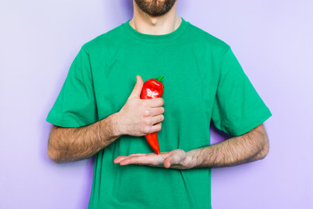 Young man holding red capi pepper in his hands showing thumb up on green t-shirt background. Light purple wall on background, copy space.の写真素材