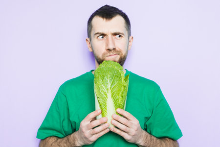 Young man holding Beijing napa cabbage in his hands, doubt face expression. Light purple background.の写真素材