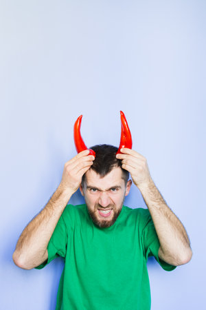 Young man holding red capi peppers above his head which looks like horns, angry and evil face. Light...