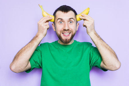 Young bearded man holding cut in half yellow banana on the level of temples, smiling and happy face expression. Light purple background, copy space.の写真素材
