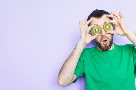 Young bearded man holding slices of green kiwi fruit in front of his eyes, surprised and shocked. Light purple background, copy space.の写真素材