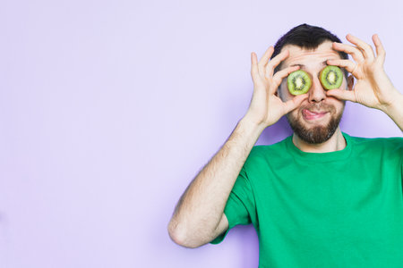 Young bearded man holding slices of green kiwi fruit in front of his eyes, showing tongue. Light purple background, copy space.の写真素材
