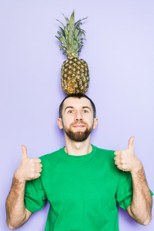 Young man holding big pineapple on his head and showing thumbs up. Light purple background, copy space, vertical orientation.の写真素材