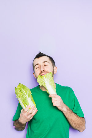 Young man biting on a leaf of Beijing napa cabbage, enjoyment face expression. Light purple background, copy space, vertical orientation.の写真素材