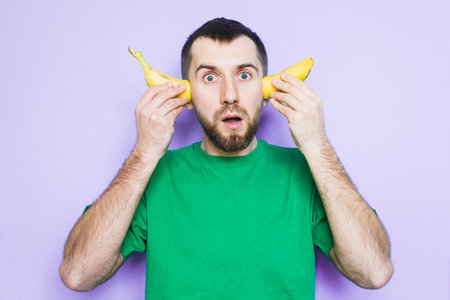 Young bearded man holding cut in half yellow banana on the level of ears, surprised and shocked face expression. Light purple background, copy space.の写真素材