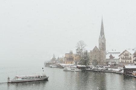 HALLSTATT, AUSTRIA - DECEMBER 2018: lake ferry approaching Evangelische Pfarrkirche and old town during snow storm.のeditorial素材