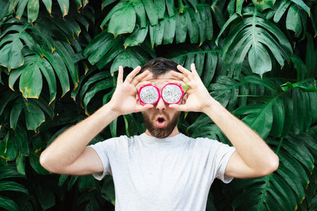 Young bearded man holding slices of Pitaya dragon fruit in front of his eyes, surprised. Tropical leaves background, copy space.の写真素材