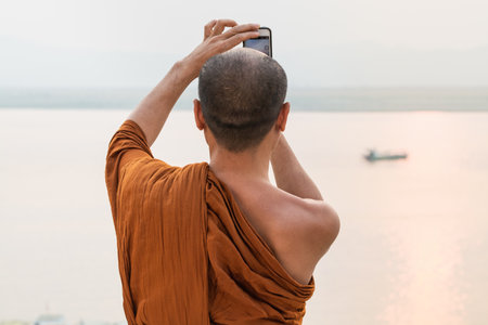 Bagan, Myanmar - March 2019: Burmese Buddhist monk in orange robe taking pictures of Irrawaddy river at sunset.のeditorial素材