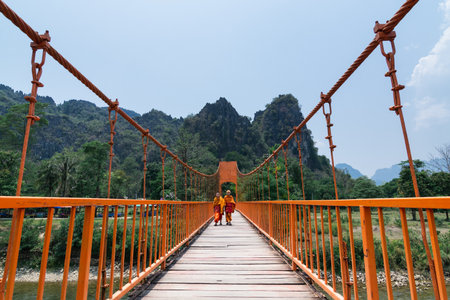 Vang Vieng, Laos - April 2019: two Buddhist monks in orange robes crossing suspension bridge.のeditorial素材