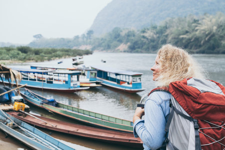 Young Caucasian blonde woman with backpack stands on boat pier in Muang Ngoi village, Laosの写真素材