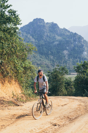 Young Caucasian man riding a mountain bicycle uphill in Muang Ngoi village, Laos. Vertical...