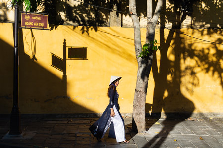Hoi An, Vietnam - June 2019: Vietnamese woman walking and dropping shadow on yellow wall in old town districtのeditorial素材