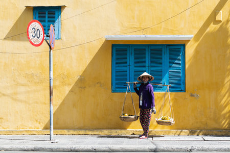Hoi An, Vietnam - June 2019: street fruit vendor woman walking by colorful house in old town.のeditorial素材