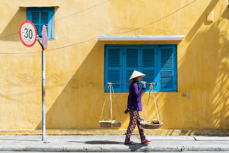 Hoi An, Vietnam - June 2019: street fruit vendor woman walking by colorful house in old town.のeditorial素材