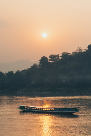 Traditional Laotian wooden slow boat on Mekong river near Luang Prabang at sunset, Laos. Golden hour, sun reflecting in waterの写真素材