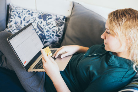 Young Caucasian woman making online purchase with credit card on the couch, looking at laptop screen...