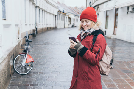Woman in red coat looking at her mobile phone screen on the street of old European townの写真素材