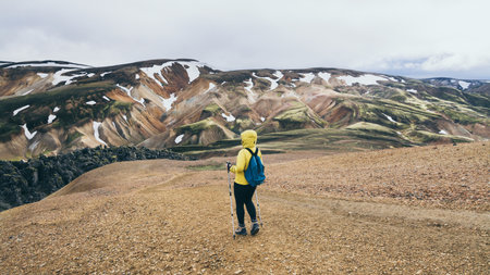 Woman in yellow raincoat hiking in the colourful mountains in Landmannalaugar national park,...