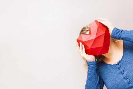 Young woman holding polygonal diamond shaped red heart in front of her head, standing in the corner.の写真素材