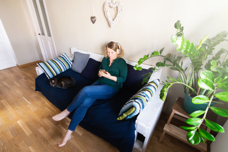 Woman listens to music in headphones on the sofa at home, holding mobile phone in her handsの写真素材
