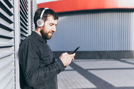 Young bearded man listening to music in wireless headphones in the urban environment, holding mobile phone in his handsの写真素材