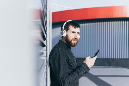 Young bearded man listening to music in wireless headphones in the urban environment, holding mobile phone in his handsの写真素材