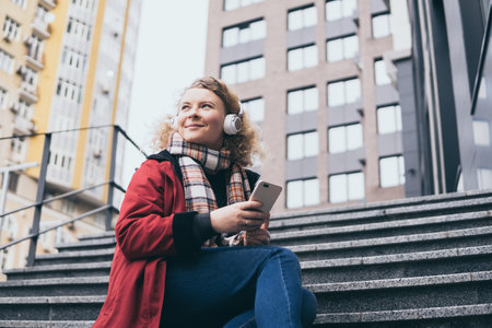 Young blonde woman listening to music in wireless headphones in the urban environment, holding mobile phone in her handsの写真素材