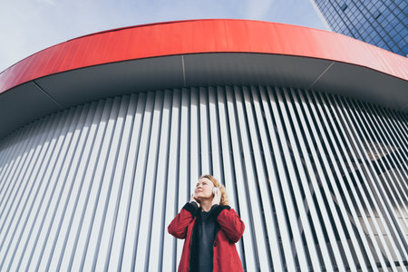 Young blonde woman listening to music in wireless headphones in the urban environment, modern architecture on backgroundの写真素材