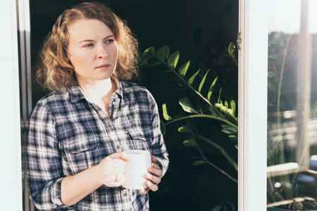 Young blonde woman looking out of the window with a concerned expression on her face.の写真素材