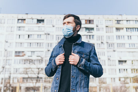 Concerned young man wearing medical mask outdoors with residential building on background. Corona virus quarantine conceptual image.の写真素材