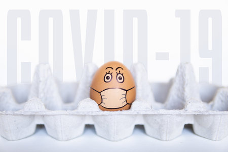 Chicken egg looking out of a tray with doodle face wearing medical mask. Conceptual image of stay at home during Corona virus quarantine, the word COVID-19 on backgroundの写真素材