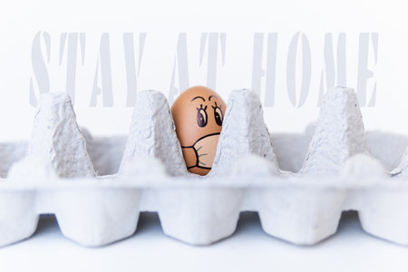 Chicken egg looking out of a tray with doodle face wearing medical mask. Conceptual image of stay at home during Corona virus quarantineの写真素材