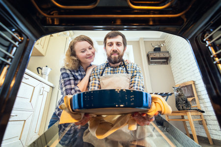 Smiling Eastern European couple cooking at home. Man taking out a pie from the oven with kitchen interior and a cat on background, view from inside the oven.の写真素材