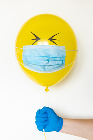Hand in a rubber glove holding a yellow balloon with a medical mask and doodle closed eyes. Conceptual image of Birthday party during Corona virus quarantine lockdownの写真素材