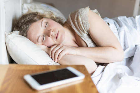 Young Eastern European woman sleeping in bed with mobile phone on the foreground.の写真素材