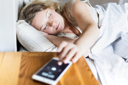 Young Eastern European woman lying in bed and turning off the alarm clock on her mobile phone in the morning.の写真素材