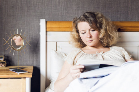 Young Eastern European woman reading a book while lying in bed in the morning.の写真素材