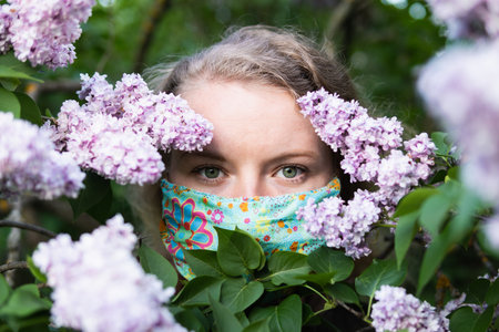 Young blonde woman smells lilac tree blossom through facial mask. Conceptual image of coronavirus quarantine and allergy.の写真素材