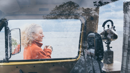 Young blond woman standing next to camper van overlooking the frozen winter sea. View through the open door.の写真素材